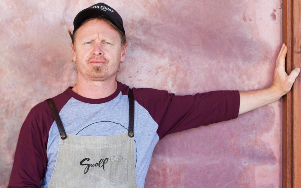 Man resting on wall of cafe wearing Swell branded apron.