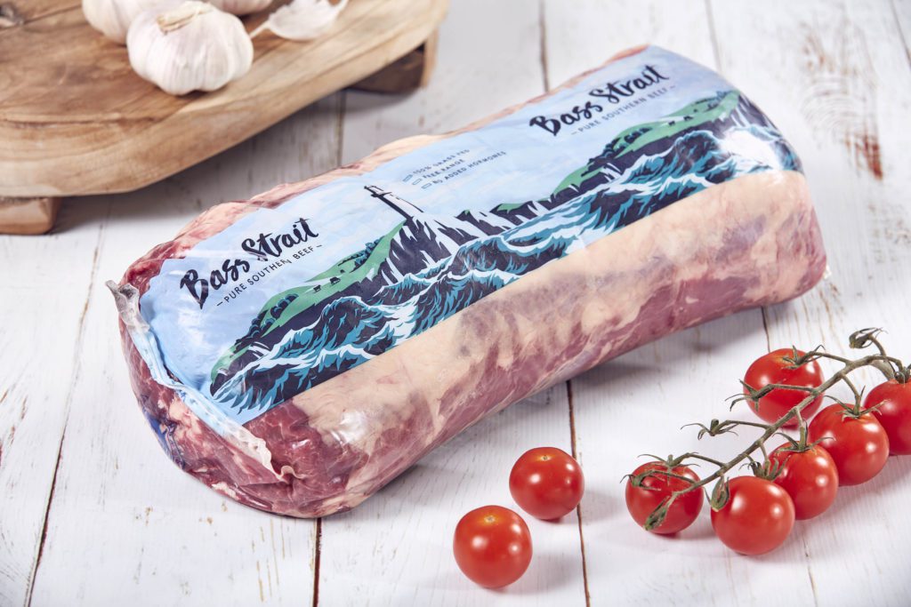 Bass Strait branding on beef product resting on a wooden surface next to vine cherry tomatoes and garlic.